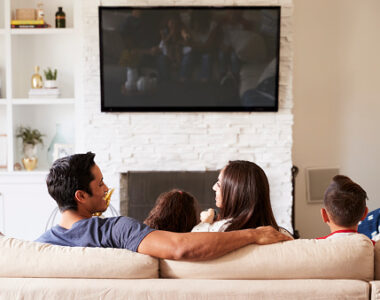 Back view of young Hispanic family of four sitting on the sofa watching TV, mum looking at dad